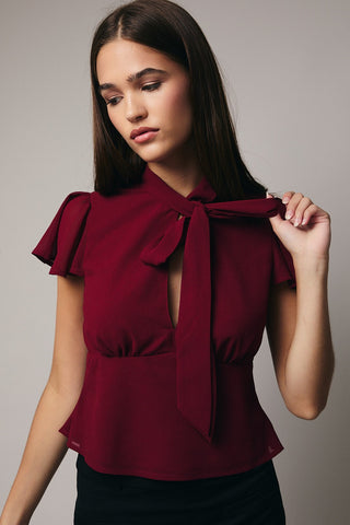 Woman wearing a burgundy blouse with ruffled sleeves and a bow tie on a gray background